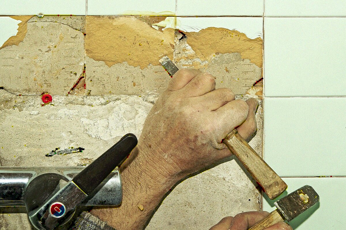 A hand using a chisel to remove wall tile and expose the substrate behind it